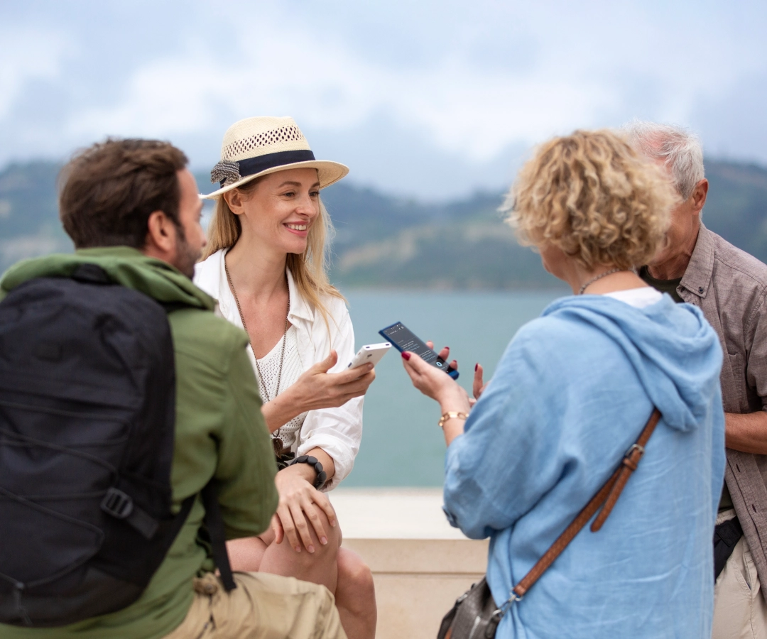 Four adults, three visible and one partially obscured, enjoy a conversation outdoors by the water. Two women, one smiling and wearing a hat, show something on their translators which they hold in their hands to the group.