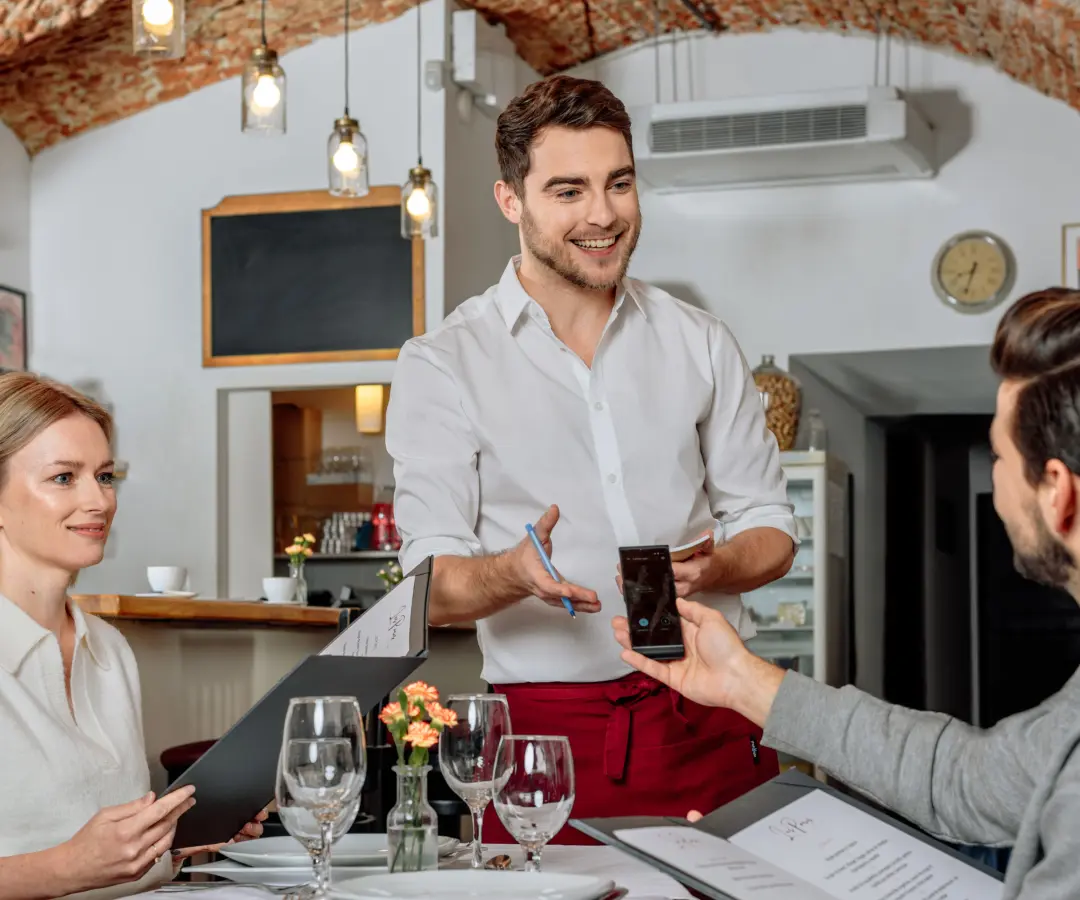 A smiling waiter stands at a restaurant table, while a seated male customer holds translator in his hand. A female customer sits nearby, holding a menu. The table is set with glasses, plates, and a small flower vase.