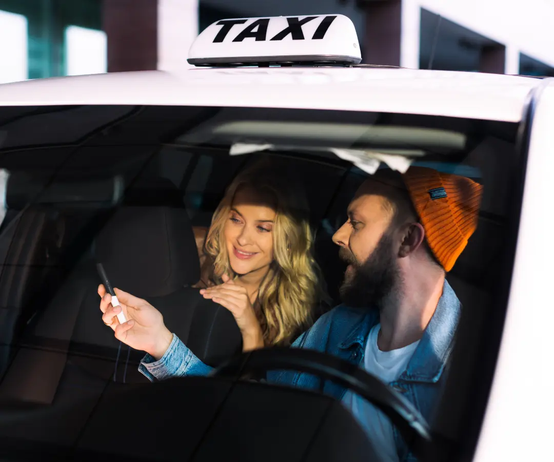 A man sits in the driver's seat of a taxi and shows something to a woman in the back seat on a translator he's holding in his hand. The woman smiles with interest. A taxi sign is visible on the roof of the car.
