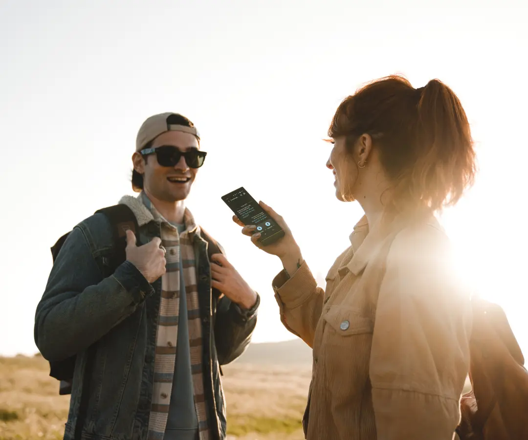 A man and woman stand in a sunny field, engaged in a lively conversation using an electronic translator. 