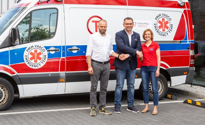 Three people stand smiling in front of a white, red, and blue ambulance parked on a paved street beside a building. Two men, one in a suit and one in a white shirt, shake hands, joined by a woman in a red shirt.