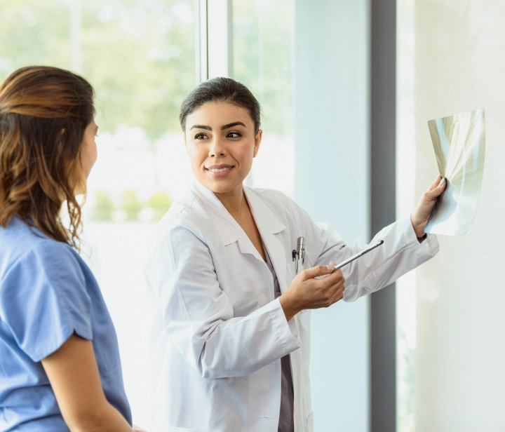 A doctor in a healthcare setting stands by a window, holding up an X-ray and explaining it to a patient.