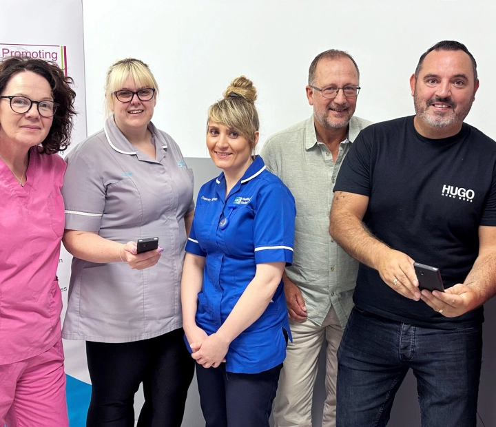 Five smiling people are standing together in healthcare attire, with two of them holding a translator device.