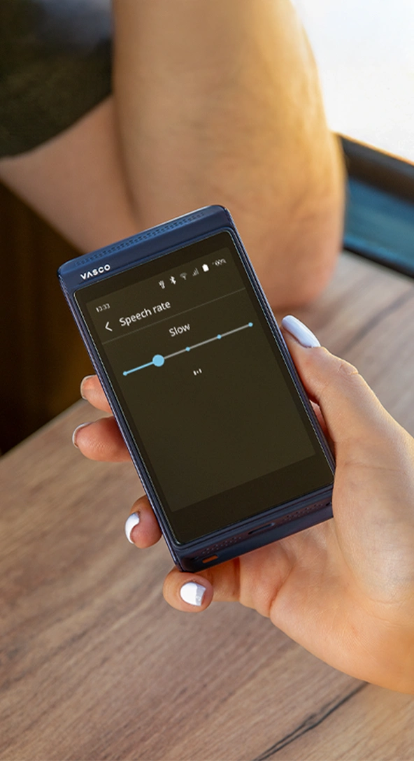 Une main tient un appareil électronique portable affichant un menu de paramètres intitulé « Voix préférées », avec des options pour la voix de l’utilisateur, la voix de l’interlocuteur et la voix du système. La personne porte un vêtement texturé jaune et blanc.