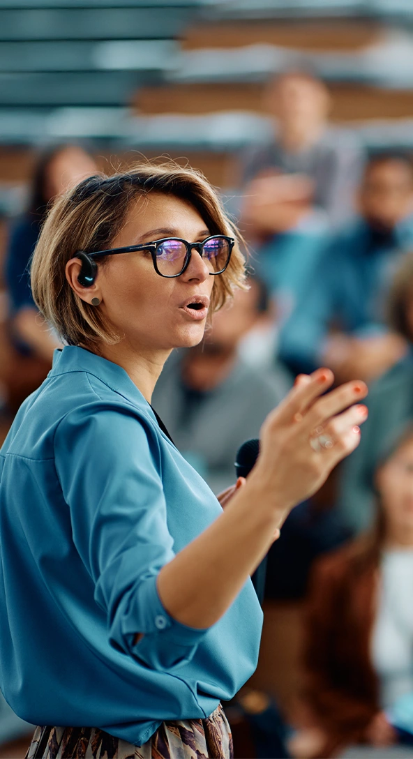 Une femme portant un casque d’écoute Vasco Translator E1 prend la parole lors d’une conférence, avec un public visible en arrière-plan.