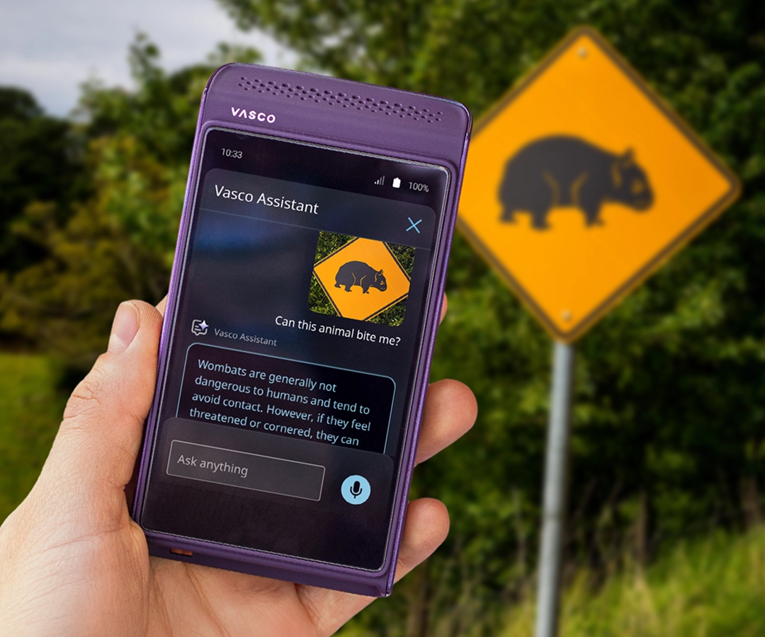 A hand holding a purple Vasco electronic translator. The Vasco Assistant app is visible on the device's screen, showing a photo of a warning sign with an image of a wombat and a description about these animals. In the background, behind the hand holding the device, is a road and the same yellow road sign with a wombat, which appears on the translator's screen.